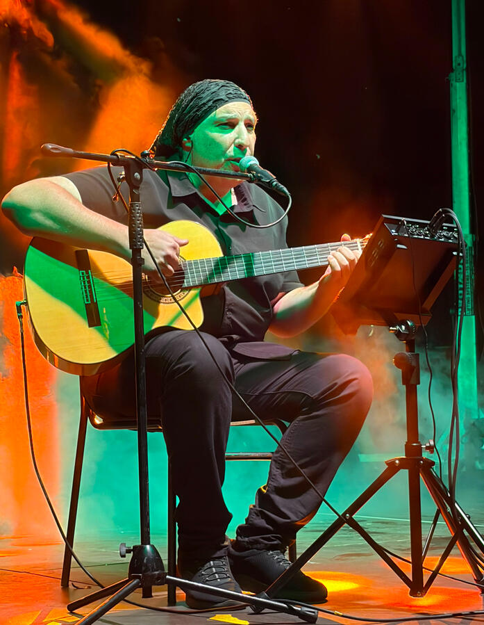 Peppone | Interprete e Cantante Peppone Ciauda che canta davanti a un microfono su un palco, mentre suona una chitarra acustica. Il musicista indossa un copricapo scuro e una camicia scura. È ripreso di profilo con una mano alzata in un gesto espressivo; l'illuminazione di scena crea un